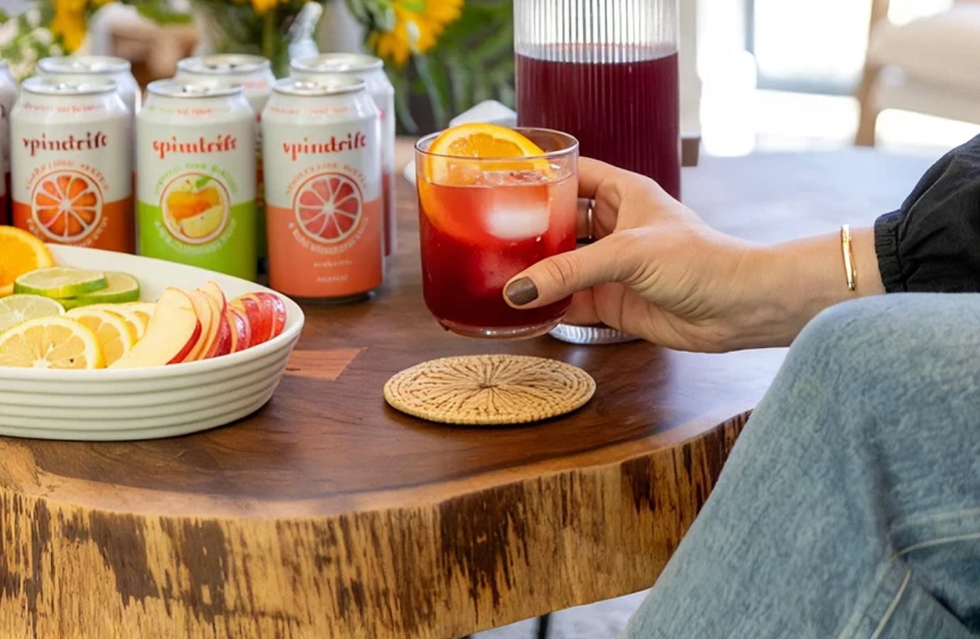 Spindrift sparkling water cans next to a glass of iced fruit drink and fresh citrus on a wooden table