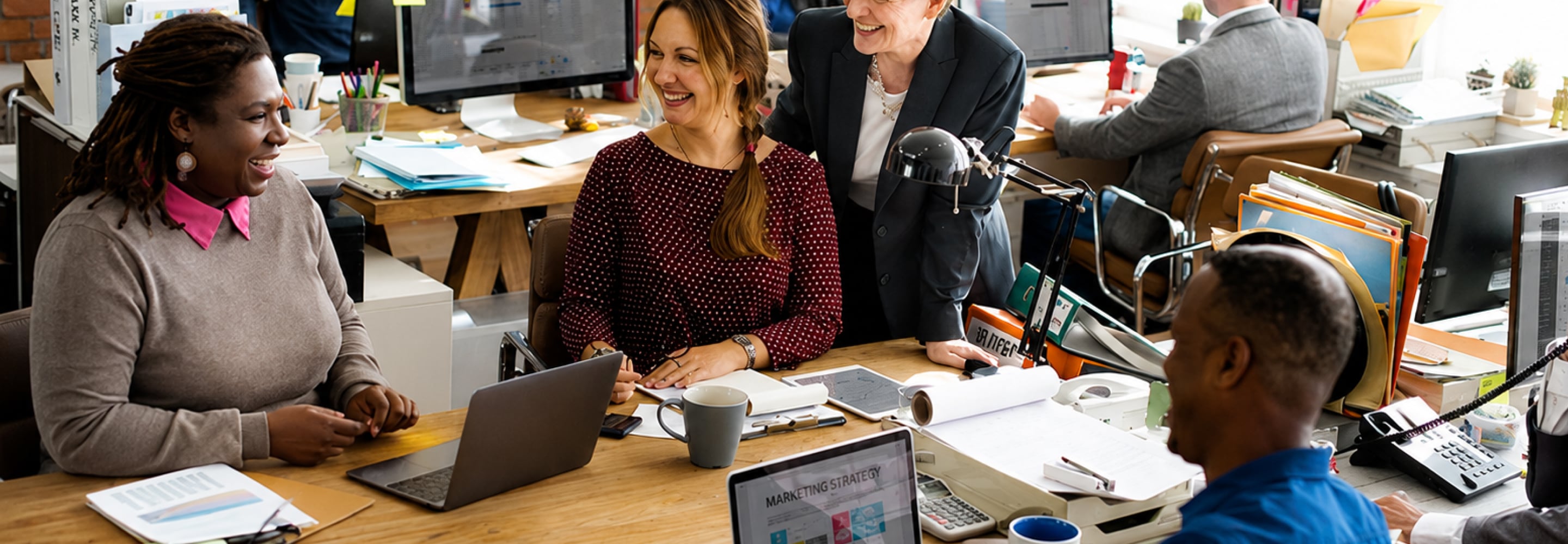 Marketing team collaborating around a table with laptops in a busy office