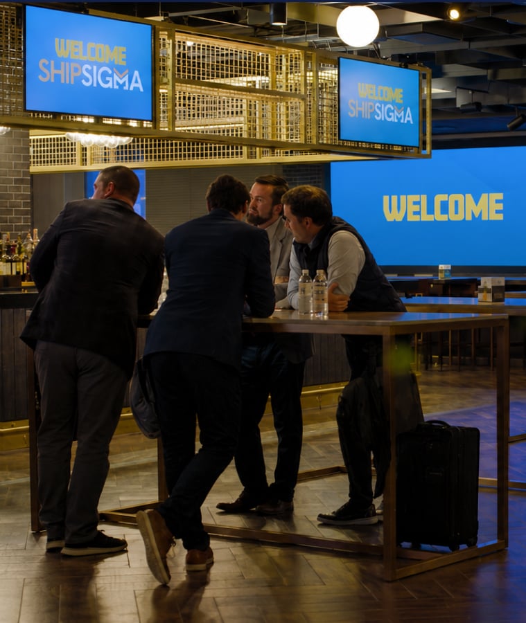 Professionals networking at a ShipSigma event with a welcome screen in the background