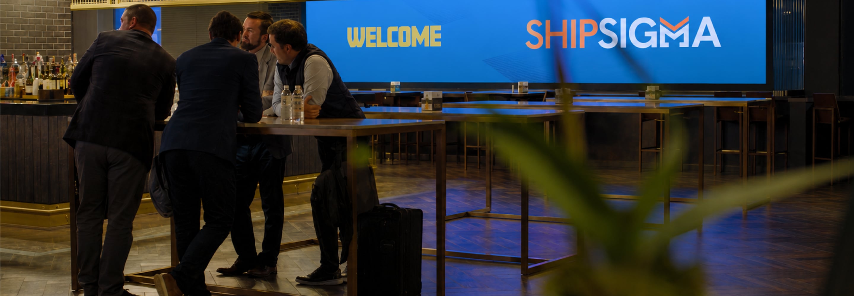 Professionals networking at a ShipSigma event with a welcome screen in the background