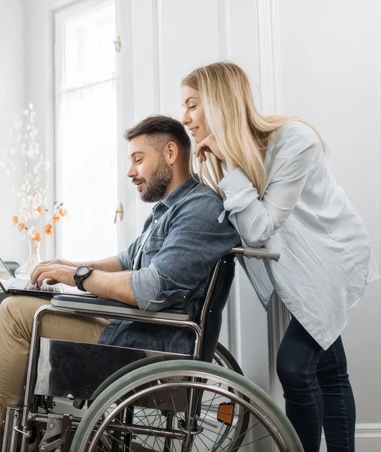 Man working on a laptop while seated in a wheelchair, with a woman beside him at home
