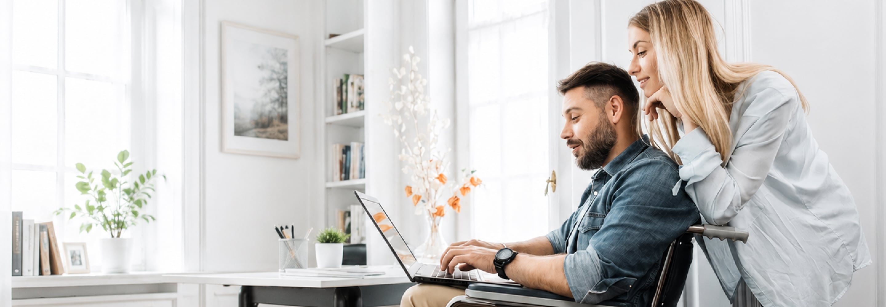 Man working on a laptop while seated in a wheelchair, with a woman beside him at home